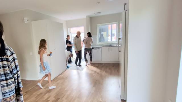 Flera personer står och pratar i ett ljust, omöblerat kök med trägolv och vita skåp; en flicka går förbi med en drink i handen.