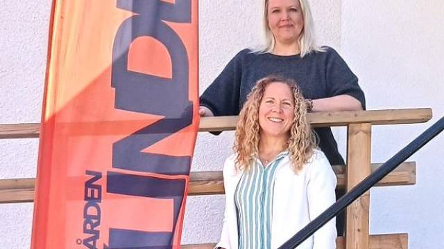 Två kvinnor står på en trätrappa bredvid en orange vertikal banner med svart text mot en vit vägg.