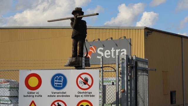 En metallstaty av en arbetare med en planka står på en grind till ett industriområde, ovanför en skylt med olika varnings- och förbudssymboler på svenska.