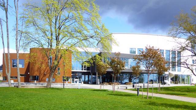 En modern byggnad med stora fönster och en brun och vit fasad, omgiven av grönt gräs, träd och en molnig himmel.