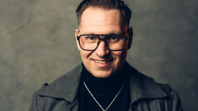 En man med kort hår och stora glasögon bär en grå kappa, svart polotröja och ett hängande halsband och står mot en neutral grå bakgrund.