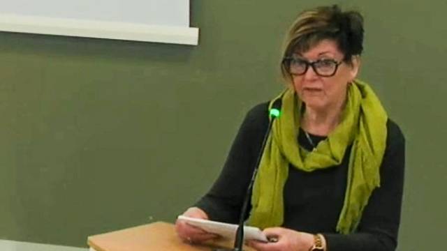 En kvinna med kort hår och glasögon talar i en mikrofon med papper i handen, sittande på ett podium framför en grön vägg.