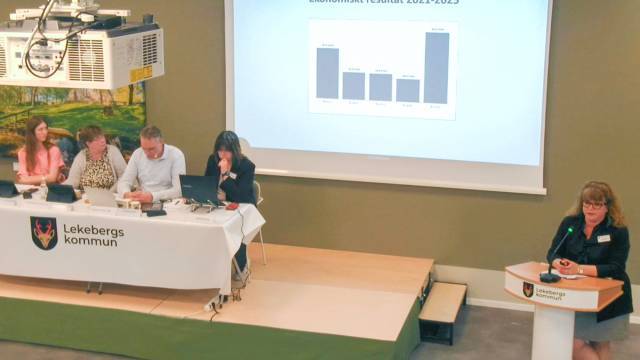 En kvinna talar vid ett podium medan fyra personer sitter vid ett bord med bärbara datorer; ett stapeldiagram projiceras på en skärm bakom dem i ett konferensrum.