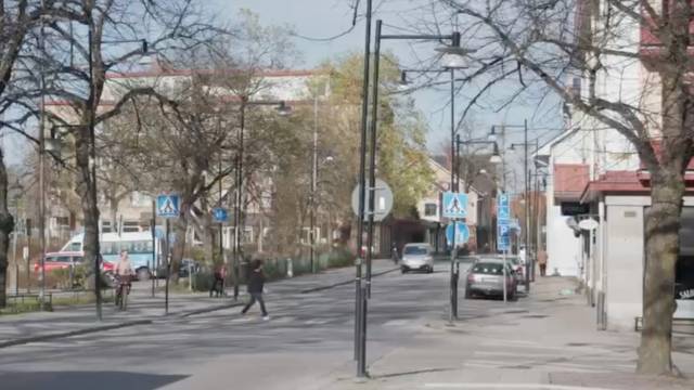 En stadsgata med fotgängare som korsar gatan, bilar parkerade längs sidorna, lövlösa träd och trafikskyltar som syns en klar dag.