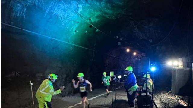 Löpare med nummerlappar och skyddshjälmar deltar i ett lopp inne i en svagt upplyst underjordisk tunnel, där evenemangspersonal och belysningsutrustning syns.