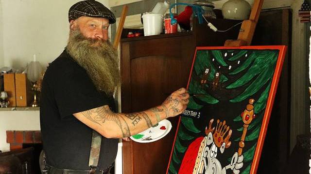 En skäggig man med tatueringar målar ett färgstarkt konstverk i tecknad stil på en duk i ett rum fyllt med hushållsartiklar.