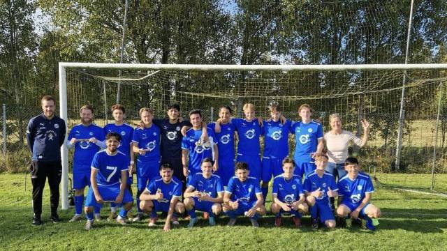 Ett fotbollslag i blå uniformer poserar tillsammans framför ett mål på en gräsplan med träd i bakgrunden.