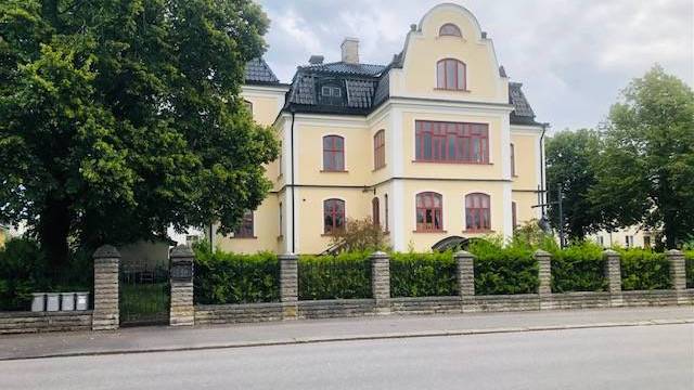 En stor gul byggnad med rödmålade fönster står bakom ett stengärde och gröna träd en molnig dag.
