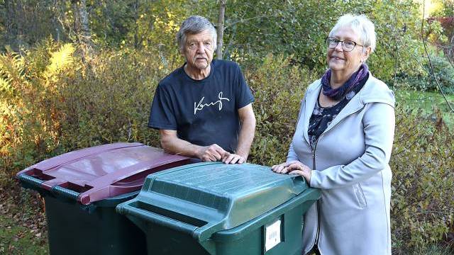 En äldre man och en kvinna står utomhus bredvid två stora soptunnor, en grön och en röd, omgivna av träd och buskar.