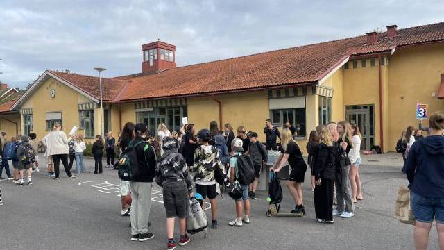 En grupp människor, däribland barn och vuxna, samlas utanför en gul byggnad med rött tak en molnig dag.