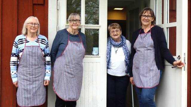 Fyra äldre kvinnor står utanför en byggnad; tre av dem bär randiga förkläden, medan en kvinna med blå halsduk står i dörröppningen.