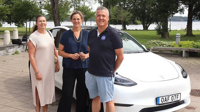 Tre vuxna står framför en vit Tesla-bil som står parkerad nära en park med träd och en sjö i bakgrunden.