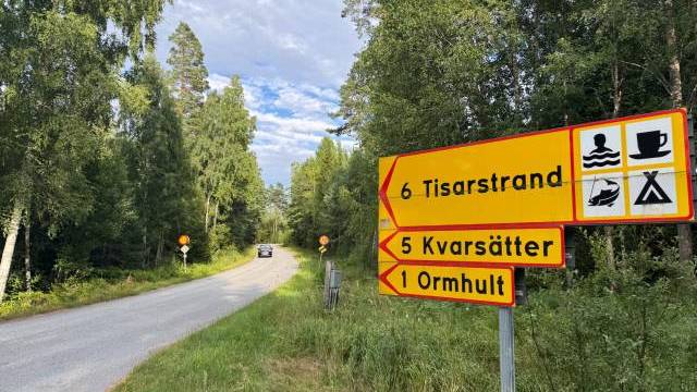 Gula vägskyltar vid en skogsväg pekar mot Tisarsstrand, Kvarsätter och Ormhult; symboler anger att det finns bad, camping och matställen i närheten. En bil kör iväg i fjärran.