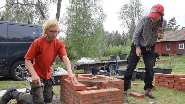 Två personer bygger en liten tegelkonstruktion utomhus; en placerar ut tegelstenar medan den andra står i närheten och tittar på. Byggverktyg och material ligger utspridda runt omkring.