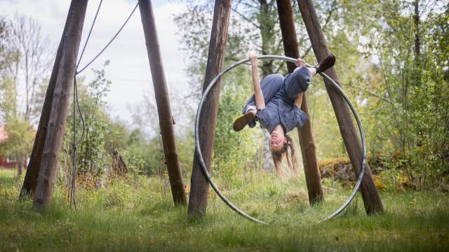 En gråklädd person hänger upp och ner i en metallring utomhus, omgiven av gräs och träd.