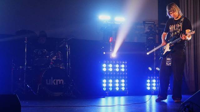 En gitarrist spelar elgitarr på en scen med blå belysning, medan en trummis är delvis synlig i bakgrunden.