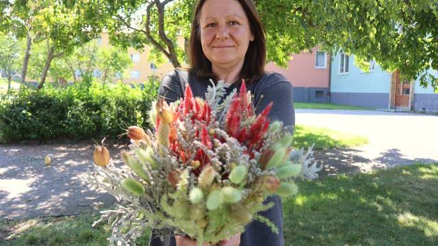 En kvinna står utomhus med en bukett bestående av olika blommor och växter i händerna, medan träd och byggnader syns i bakgrunden.