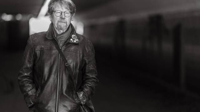 En äldre man med glasögon och skägg står i en gångtunnel, iklädd en skinnjacka och bär en axelremsväska. Bilden är i svartvitt.