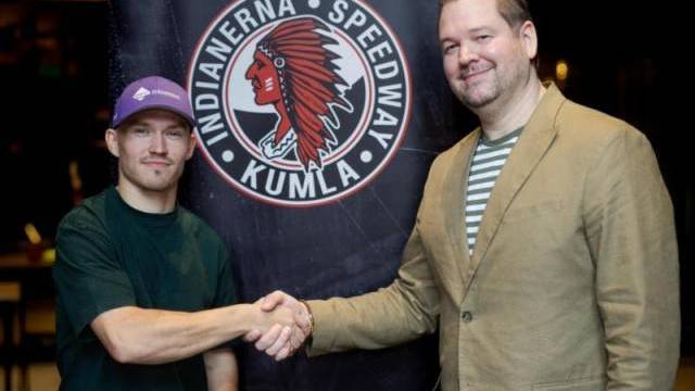 Två män skakar hand framför en Indianerna Speedway Kumla-logotyp, den ene i lila keps och vardagskläder, den andre i beige kavaj och randig skjorta.