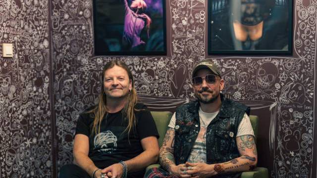 Två män med tatueringar och långt hår sitter på gröna stolar framför en vägg täckt av klotter, med två inramade foton hängande ovanför dem.