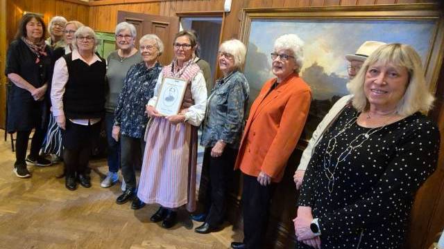 En grupp på elva äldre vuxna står inomhus i en rad, med en person i mitten som håller ett inramat certifikat. Målningar pryder de träpanelklädda väggarna bakom dem.