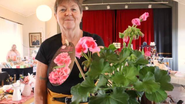En äldre kvinna som håller i en krukväxt med rosa pelargon står inomhus på en marknad eller mässa, med bord med varor och en scen med röd ridå i bakgrunden.