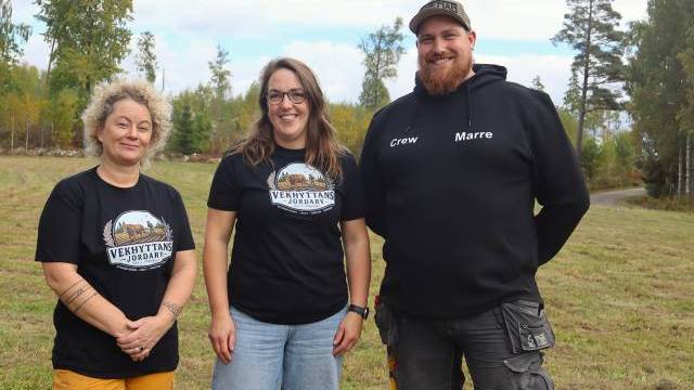 Tre personer står på en gräsbevuxen äng; två av dem bär T-tröjor med texten ”Vekhytan Nordarf” och en har en svart huvtröja med texten ”Crew Marre”. I bakgrunden syns träd och en molnig himmel.