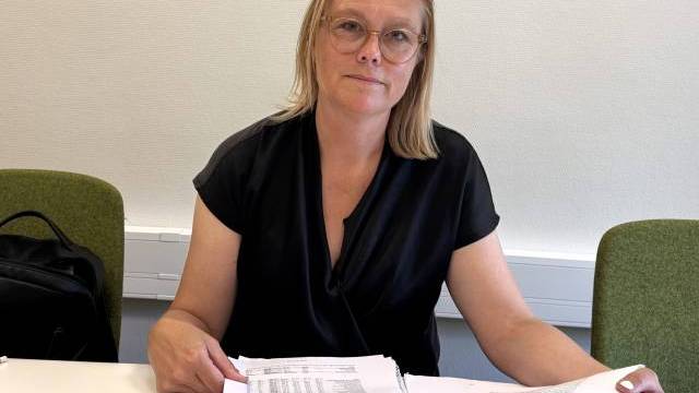 En kvinna med glasögon sitter vid ett skrivbord, håller i en bunt papper och tittar mot kameran. Hon sitter mellan två gröna stolar.