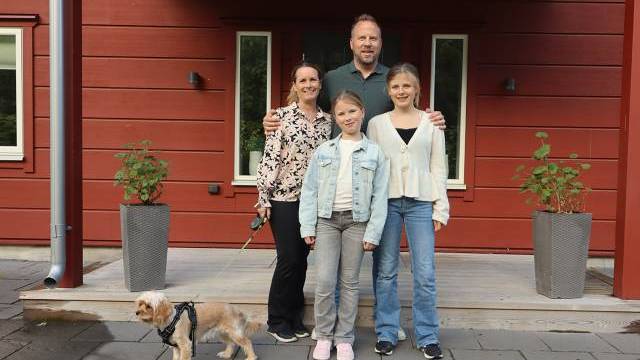 En familj på fyra personer står framför ett rött hus, med två krukväxter och en liten hund i koppel till vänster.
