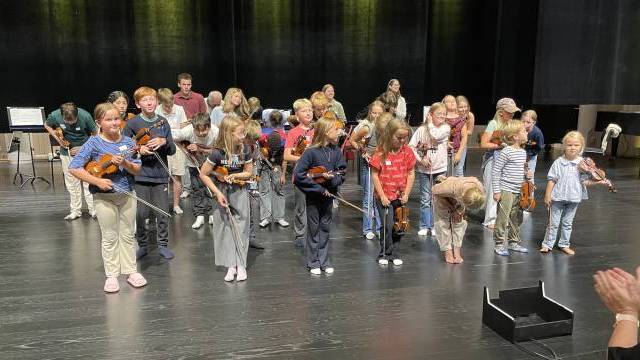 En grupp barn med violiner står på scenen, några ler och bugar, med vuxna och andra barn i bakgrunden.