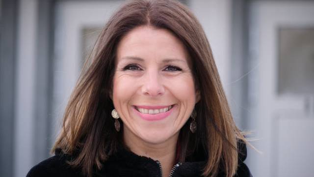 En kvinna med rakt brunt hår, klädd i svart kappa och bladformade örhängen, ler utanför en byggnad.