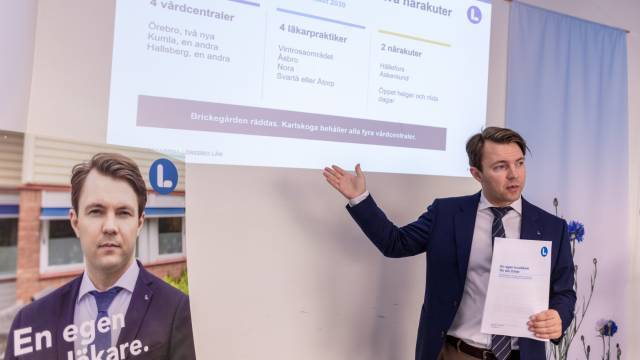 En man i kostym visar upp data på en skärm samtidigt som han håller i ett papper; bredvid honom står en affisch med hans foto och text på svenska.
