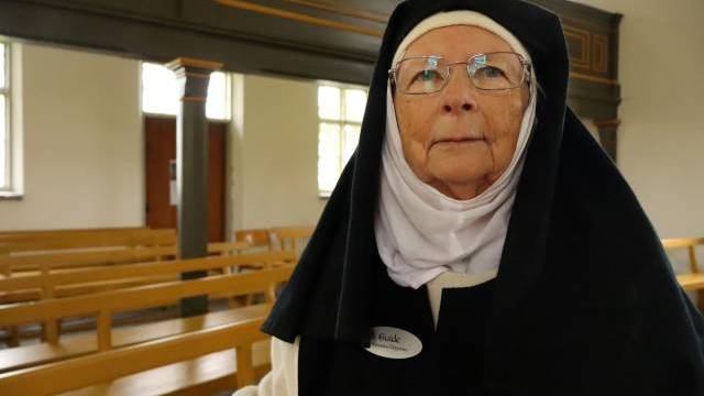 En äldre kvinna i nunnedräkt och glasögon står i en kyrka med bänkar av trä. Hon har en namnskylt fäst på sina kläder.