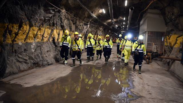 En grupp arbetare i skyddsutrustning går genom en underjordisk tunnel med steniga väggar och våta fläckar på marken.