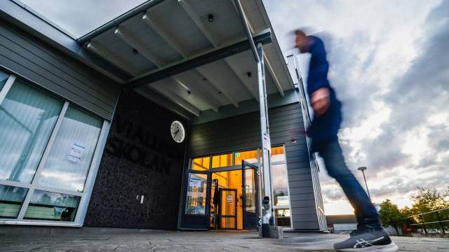 En person går förbi ingången till en modern skolbyggnad i skymningen; ovanför dörren hänger en klocka och lamporna lyser inne i byggnaden.