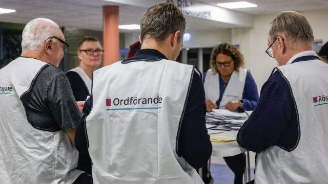 Fem personer i vita västar sitter vid ett bord och sorterar papper inomhus; de är troligen sysselsatta med rösträkning i samband med ett val eller med administrativt arbete.