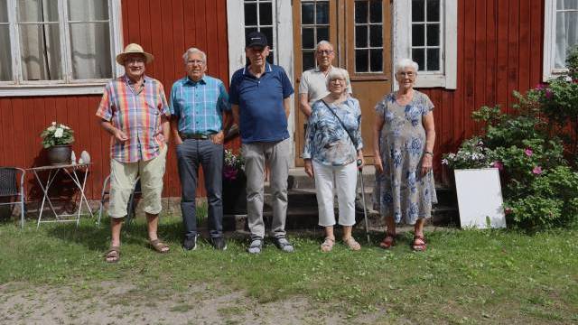 Sex äldre personer står på rad utanför en röd träbyggnad med stora fönster och en dubbeldörr, omgiven av grönt gräs och blommor.