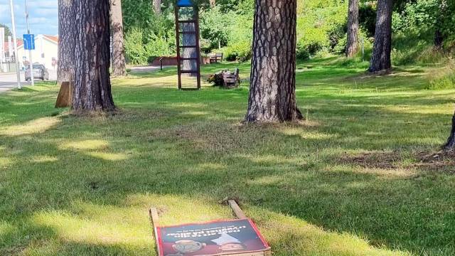 En cornhole-bräda i trä med ett illustrerat motiv står på gräset mellan träden i en park. I bakgrunden syns en byggnad med blått tak och bänkar.