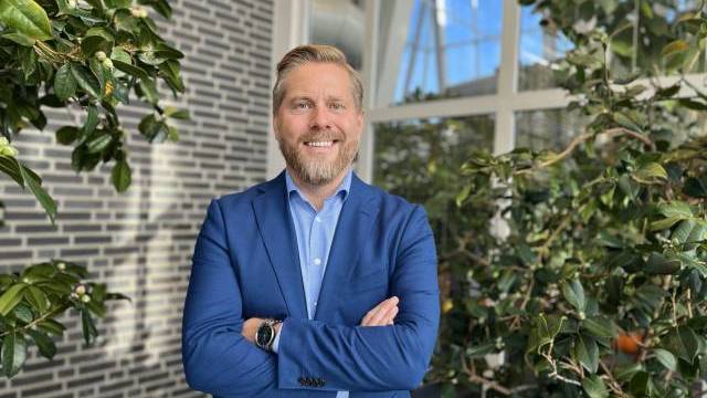 En man i blå kostym står inomhus med armarna i kors och ler, omgiven av gröna växter och stora fönster i bakgrunden.