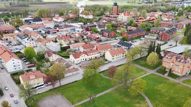 Flygbild över en liten stad med hus med röda tak, trädkantade gator, en kyrka och gröna öppna ytor en klar dag.