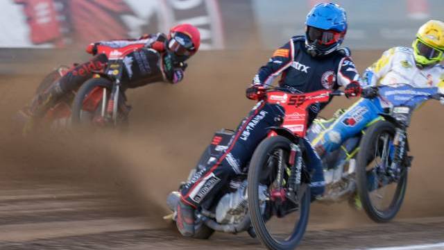 Tre motocrossförare tävlar på en grusbana och lutar sig in i en kurva medan damm virvlar upp bakom dem. I den suddiga bakgrunden syns åskådare och en stor banderoll.