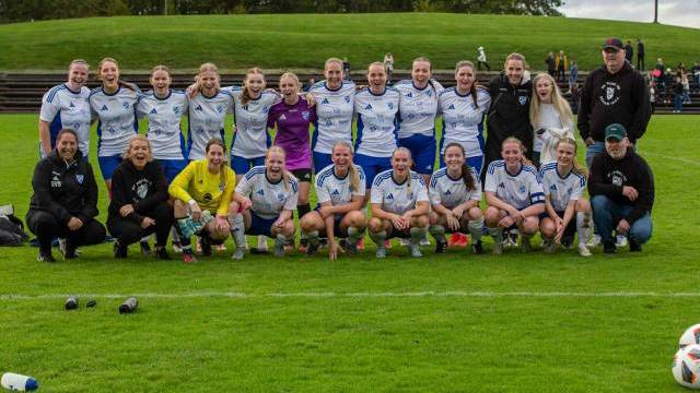 Ett damfotbollslag i vita och blå uniformer poserar för ett gruppfoto med tränare och personal på en gräsplan.