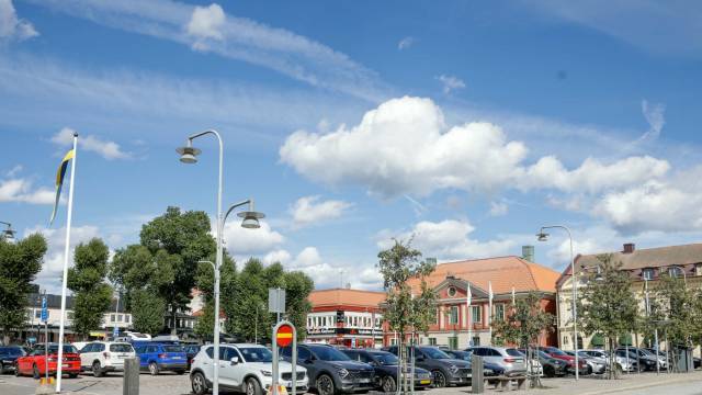 En parkeringsplats full av bilar en solig dag, med byggnader och träd i bakgrunden under en blå himmel med moln.