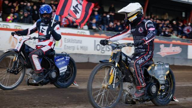Två speedwayförare i tävlingskläder tävlar på en grusbana; den ena föraren håller i en röd och svart flagga. Åskådare tittar på från läktaren i bakgrunden.
