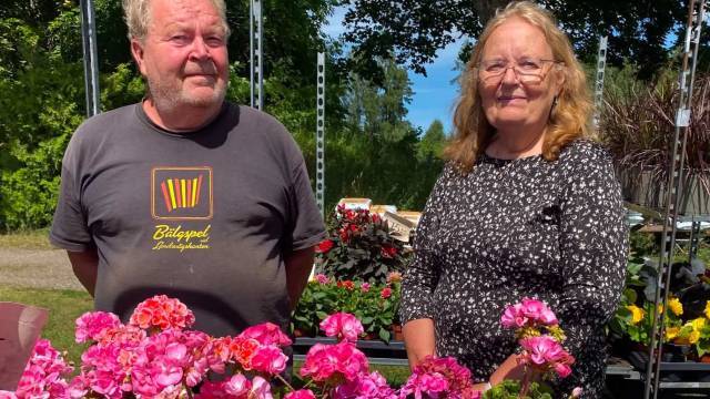 Två personer som står utomhus bakom ett bord med rosa och röda blommor, med grönska och krukväxter i bakgrunden.