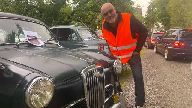 En man klädd i en knallorange säkerhetsväst står bredvid en klassisk svart bil på en grusväg kantad av andra fordon och träd.