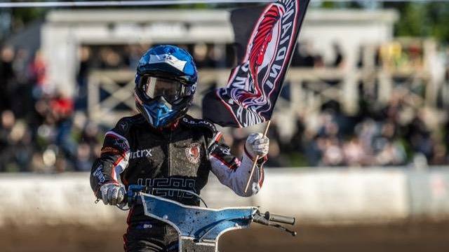 En speedwayförare i tävlingskläder och hjälm håller en lagflagga medan han kör motorcykel på en grusbana, med åskådare i bakgrunden.