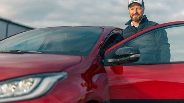 En man med keps och jacka står bredvid en öppen dörr på en röd bil och ler mot kameran under en molnig himmel.