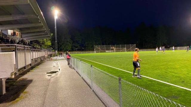 En fotbollsmatch spelas på kvällen på en upplyst plan, med en domare i orange och åskådare som sitter under en täckt läktare till vänster.