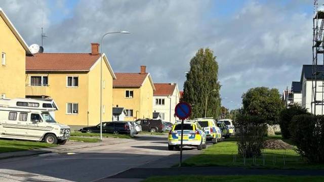 Flera polisbilar står parkerade längs en bostadsgata kantad av gula hus under en delvis molnig himmel.